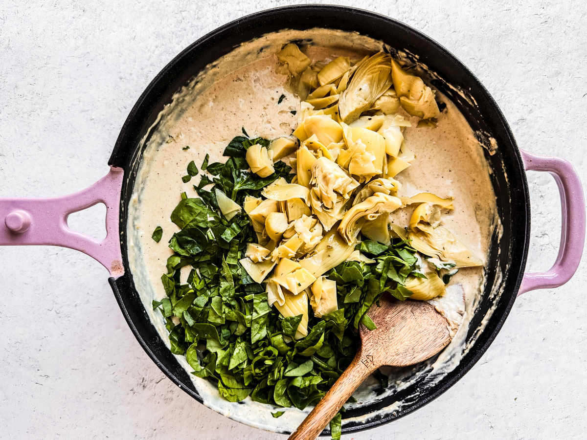 Skillet with artichokes and spinach dumped into cheese sauce.