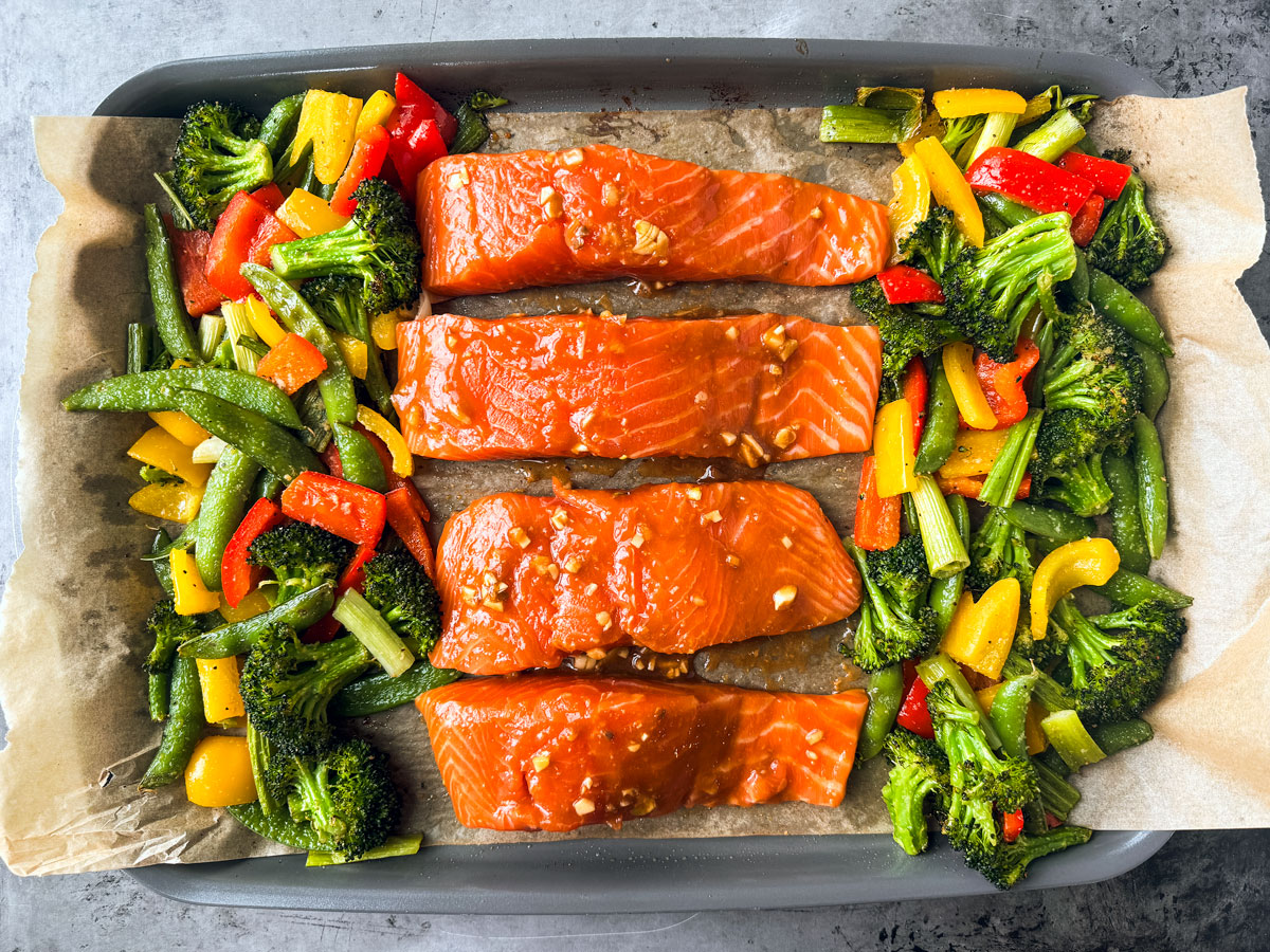Veggies and uncooked marinated salmon fillets on a baking sheet.