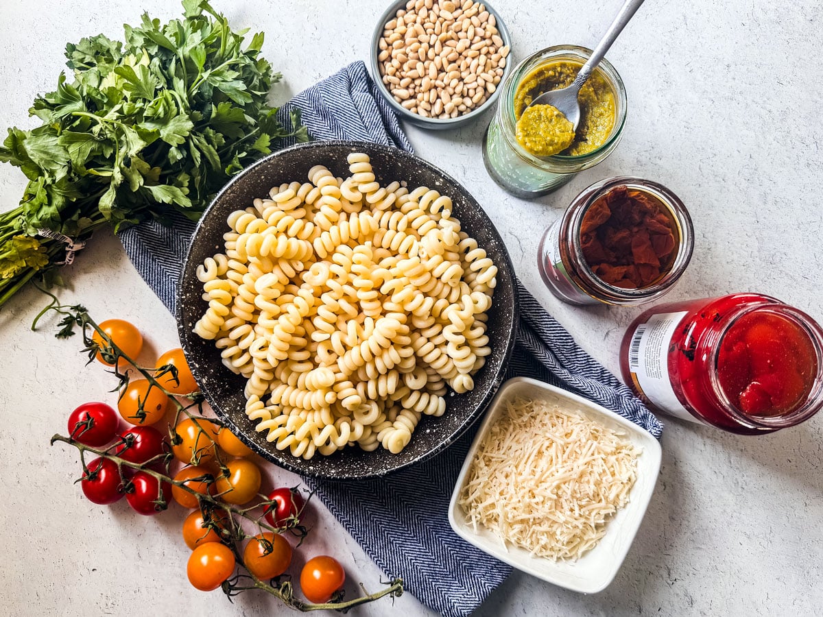 The ingredients for pesto pasta salad with sun-dried tomatoes are spread out on a white background.