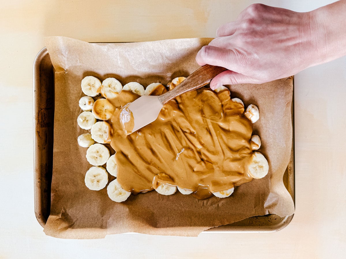 Peanut butter being spread over banana slices on a sheet pan.