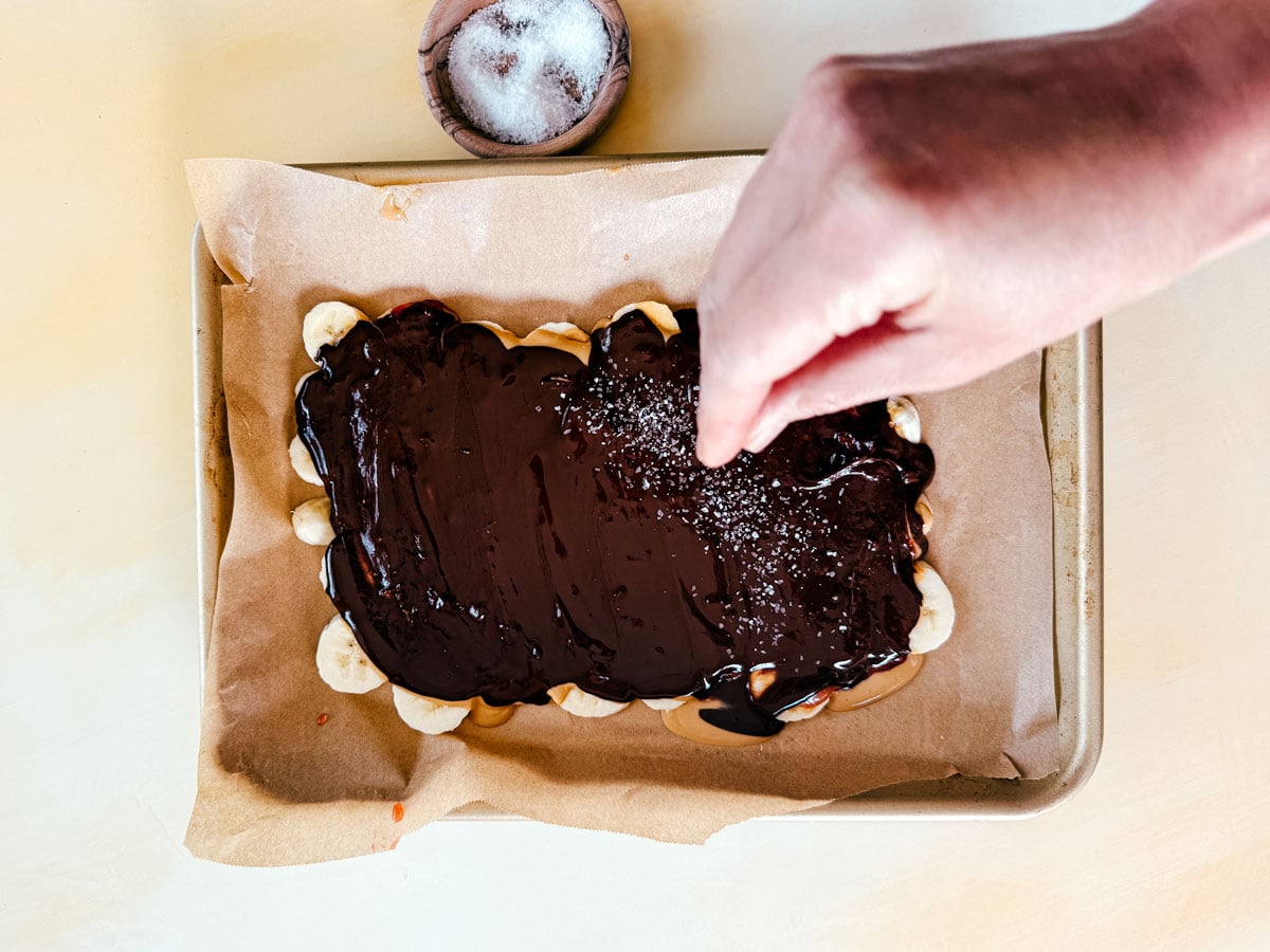 Banana slices smothered in chocolate and being sprinkled with sea salt.