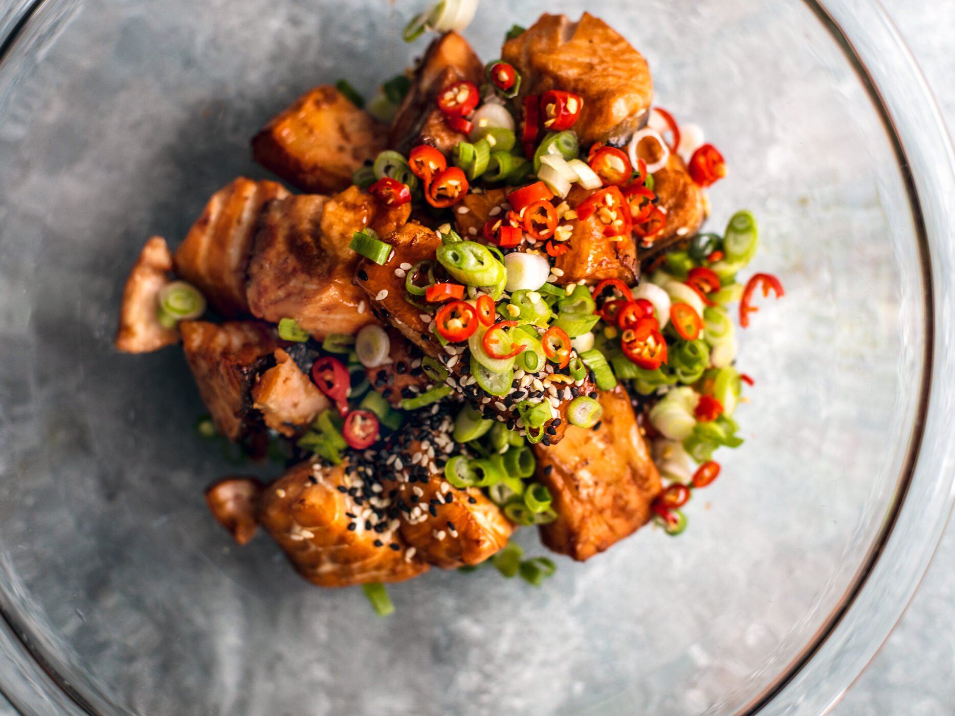 Glass mixing bowl full of salmon bites topped with sweet chili sauce, scallions, sesame seeds, and Thai chilies.