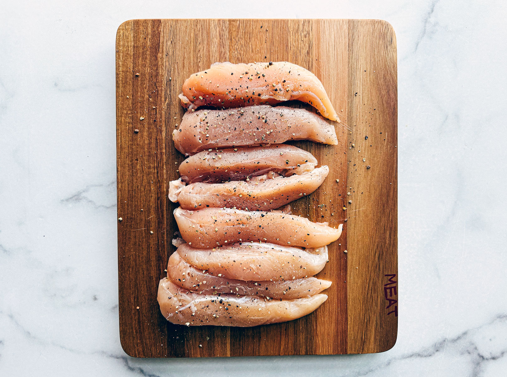 Wooden cutting board with uncooked chicken tenderloins on it.