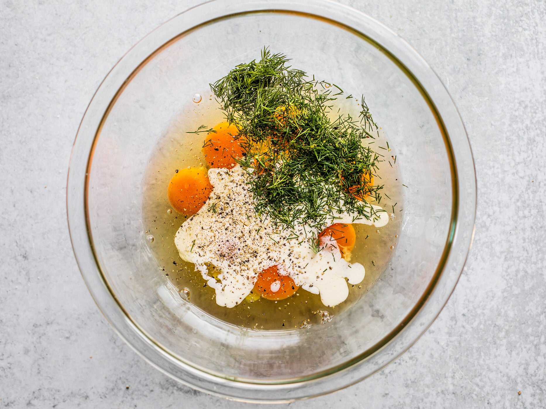Glass mixing bowl of eggs, cream, fresh chopped dill, and cracked pepper.