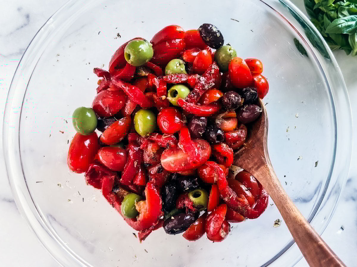 Glass mixing bowl full of seasoner of tomatoes, roasted red peppers, and olives.