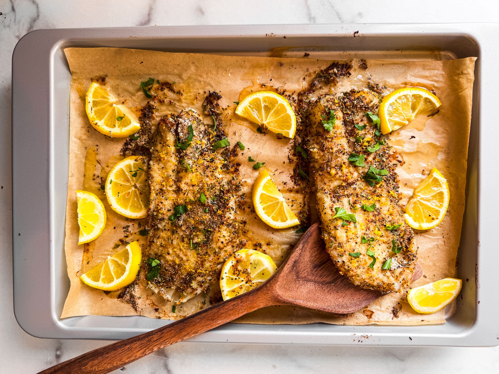 Baked lemon pepper grouper on a baking sheet with lemon wedges garnished with fresh parsley.