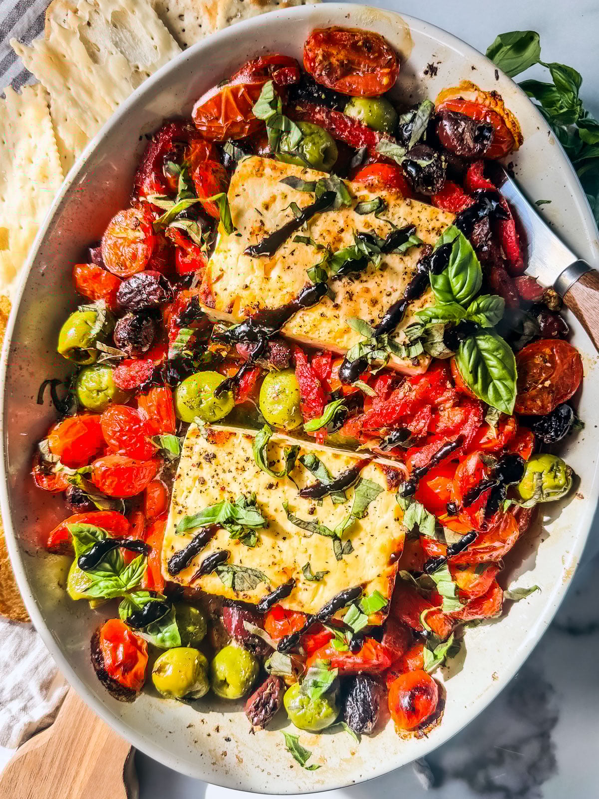 Baked feta in a dish with tomatoes, roasted red peppers, and olives garnished with basil and drizzled with balsamic glaze.