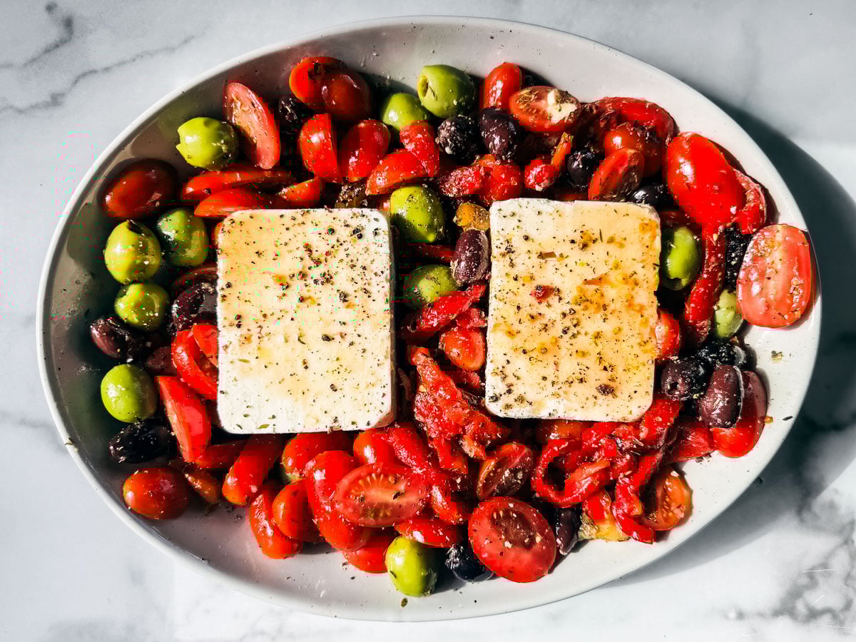 Two chunks of feta in a baking dish surrounded by tomatoes, olives, and roasted red peppers.