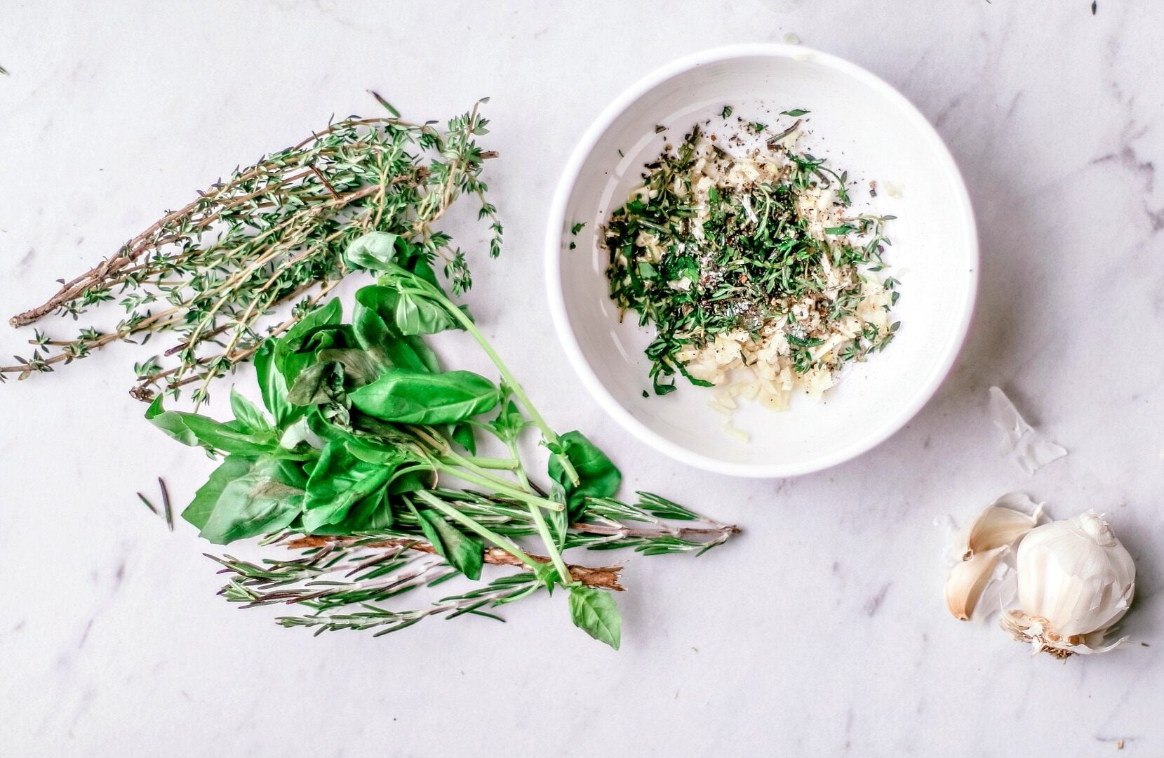 Fresh herbs and garlic next to a bowl of chopped herbs and garlic.
