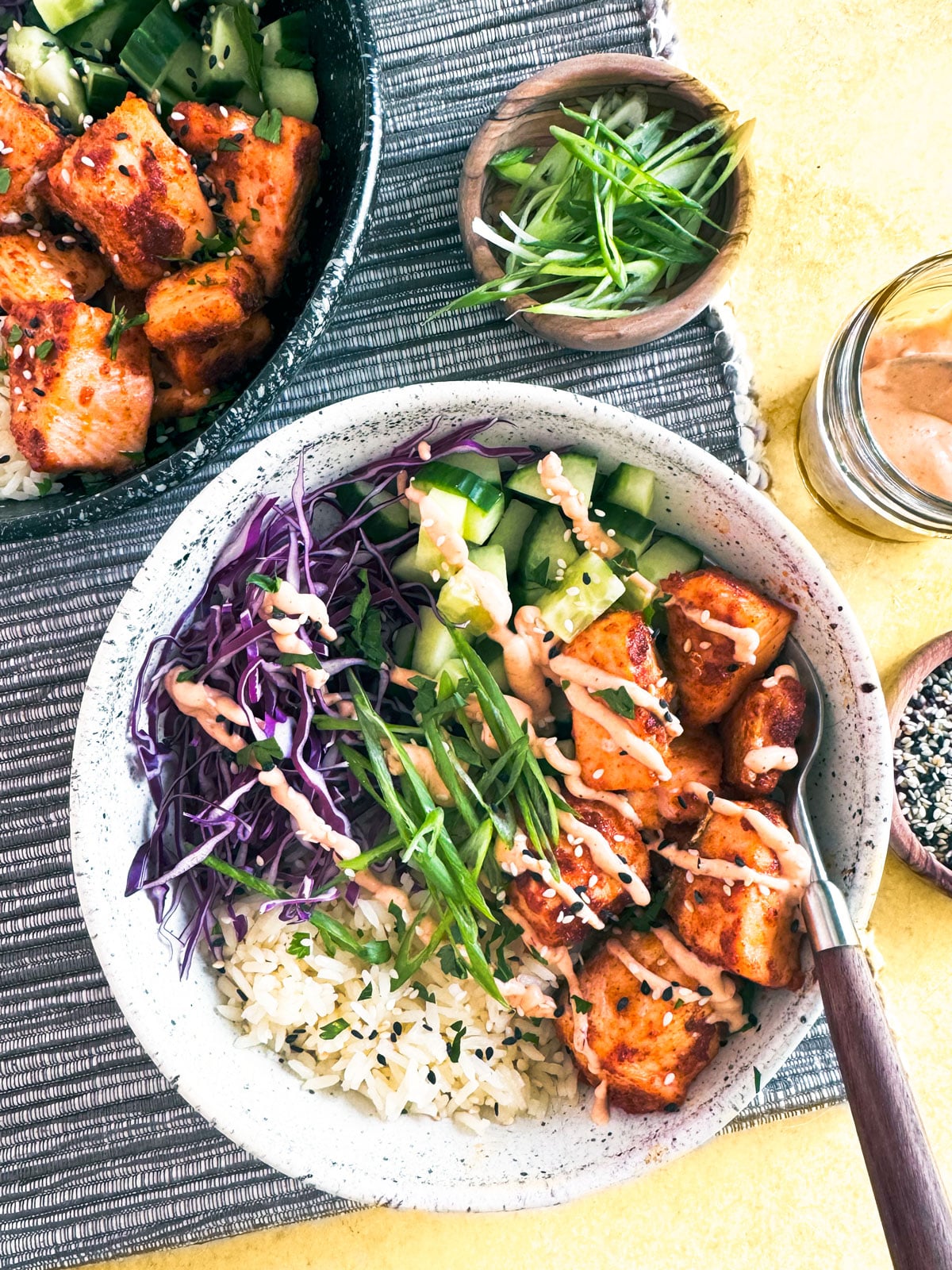 Two Bang Bang salmon bowls on a yellow surface with bang bang sauce in a jar and scallions in a pinch bowl.