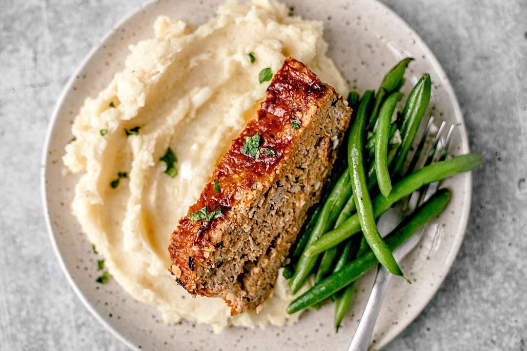 Slice of meatloaf on a plate with mashed potatoes and green beans.