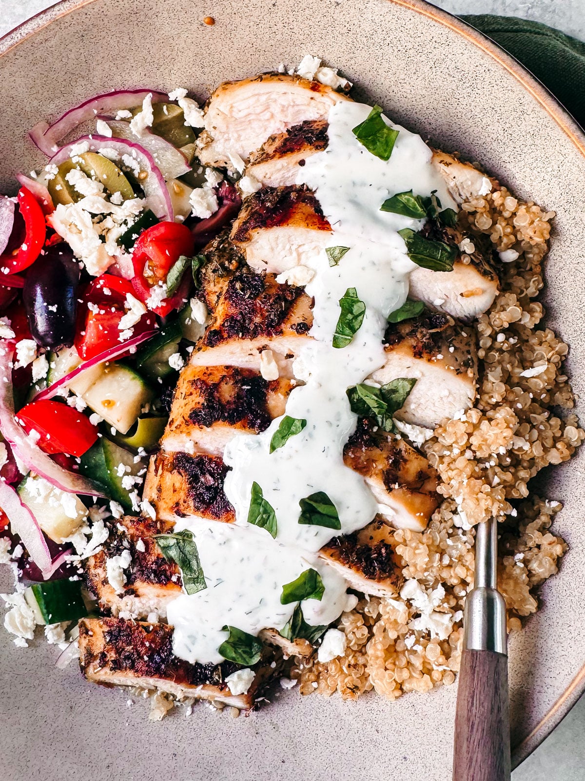 Bowl of quinoa topped with grilled sliced chicken, fresh veggies, and tzazizki.