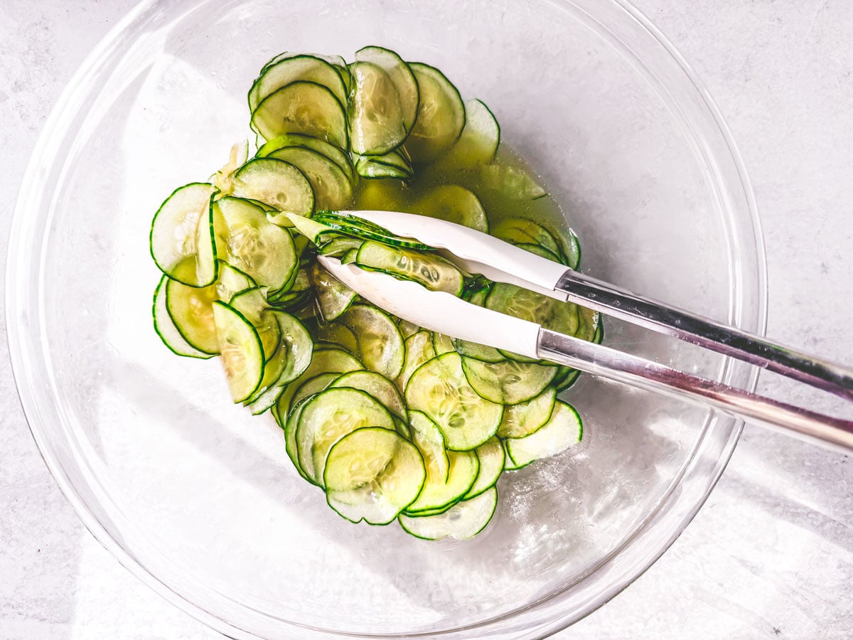 Cucumber slices being tossed in a bowl with tongs.