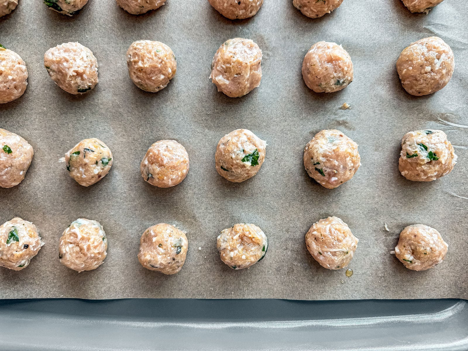Ground chicken meatball ingredients in a glass mixing bowl next to a baking sheet of uncooked chicken meatballs.